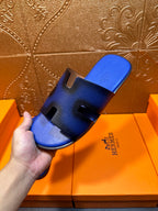 Hand holding blue textured slide sandal with black cutouts and Hermes logo on side