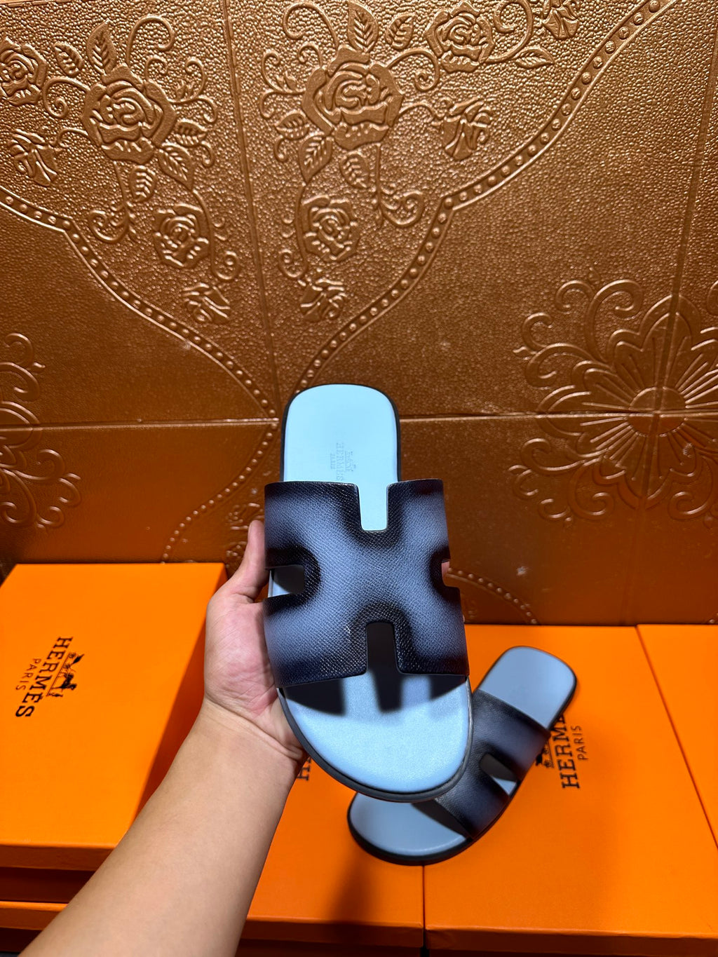 Hand holding dark navy leather slide sandals with light blue footbeds and HERMES marks.