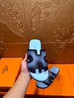 Hand holding dark navy leather slide sandals with light blue footbeds and HERMES marks.
