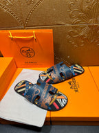 Blue Hermes logo slide sandals with patterned straps and open toe, diagonal view.