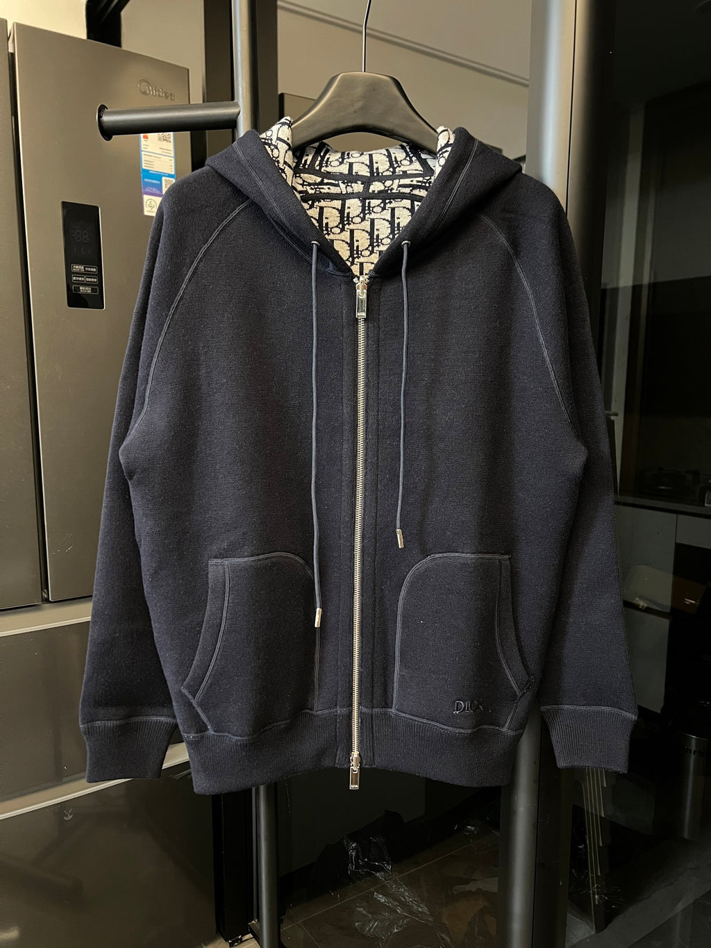 Navy zip-up hoodie with hood, front pockets, and beige Dior logo patterned lining.