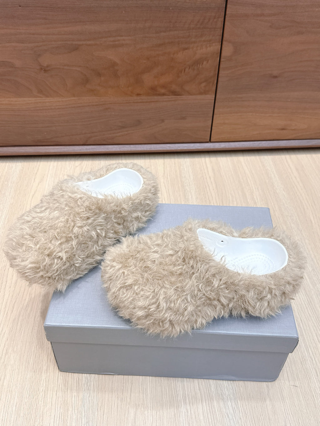 Beige faux-fur slide sandals on gray box, angled view