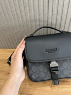 Coah crossbody bag close-up; black flap with emblem, patterned front panel, top handle strap
