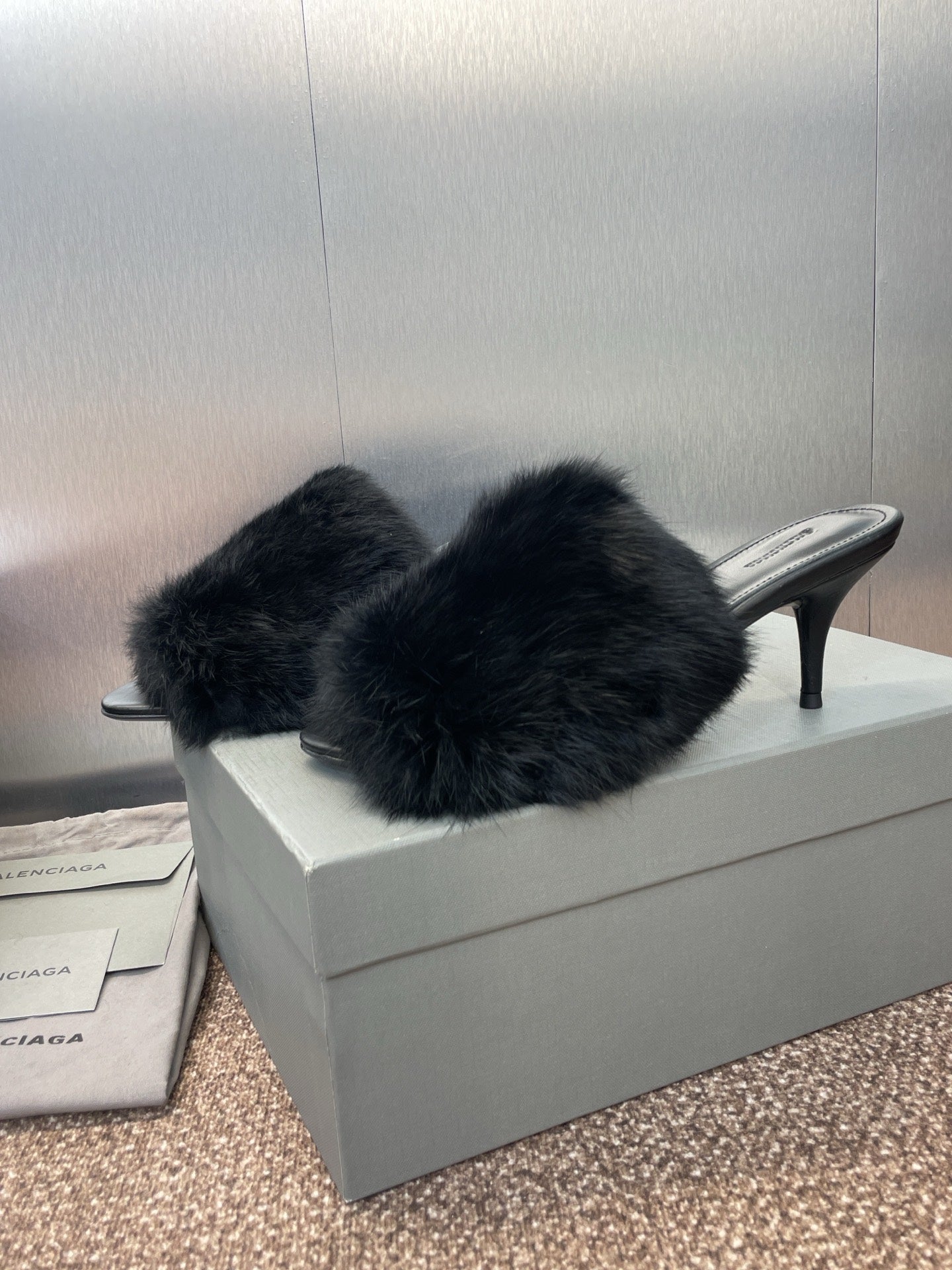 Black faux-fur slide sandals with open toe and slim high heel on box.