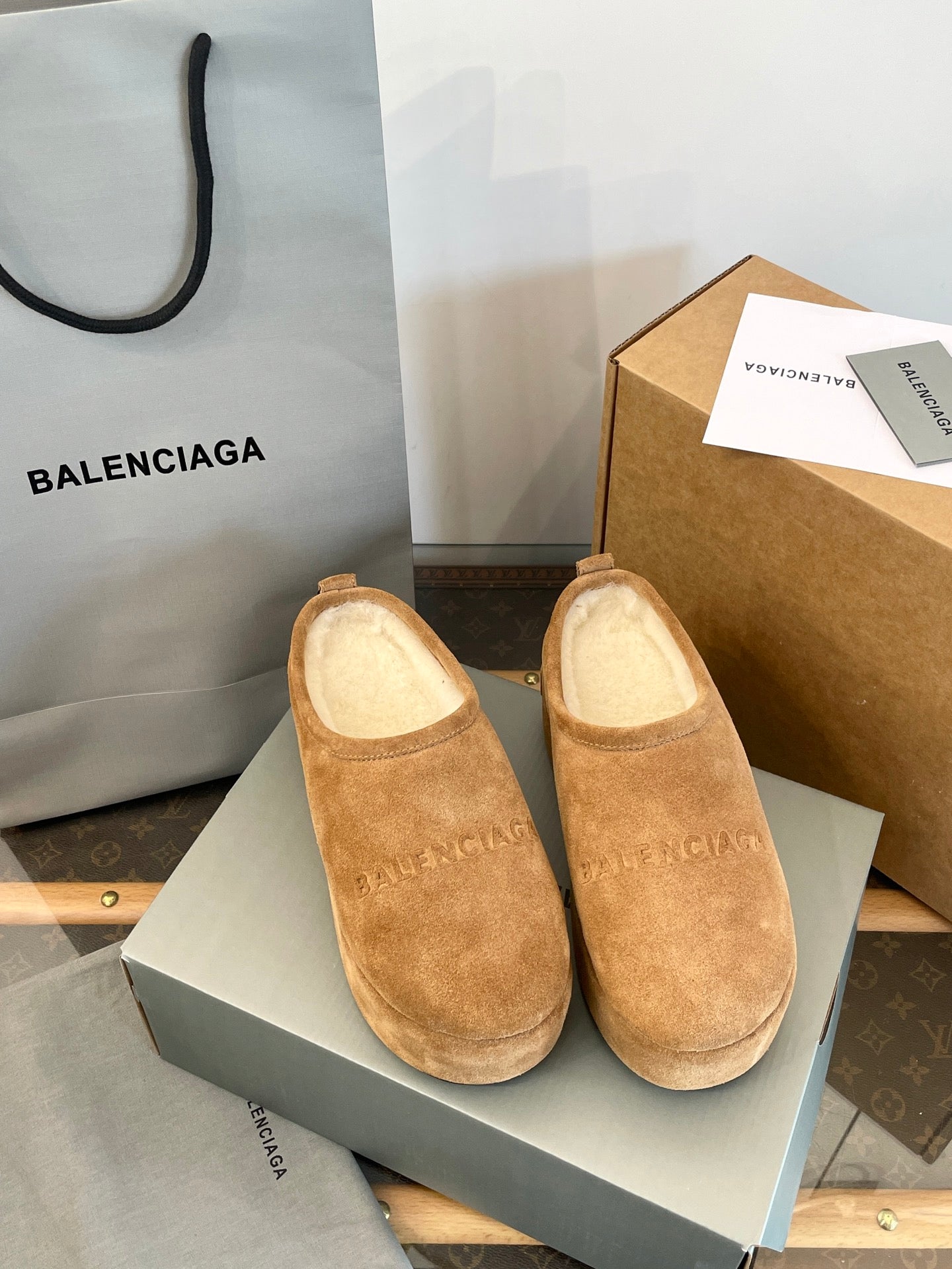 Tan suede slip-on sandals with beige lining and curved toe, shown in a shoe box.