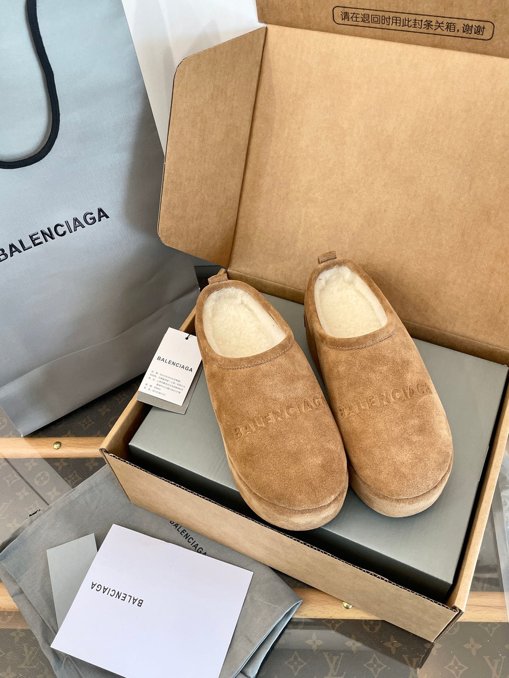 Tan suede slip-on sandals with beige lining, pair displayed in an open shoebox.