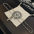 Silver-tone sword pendant necklace with floral chain links beside a cloth pouch