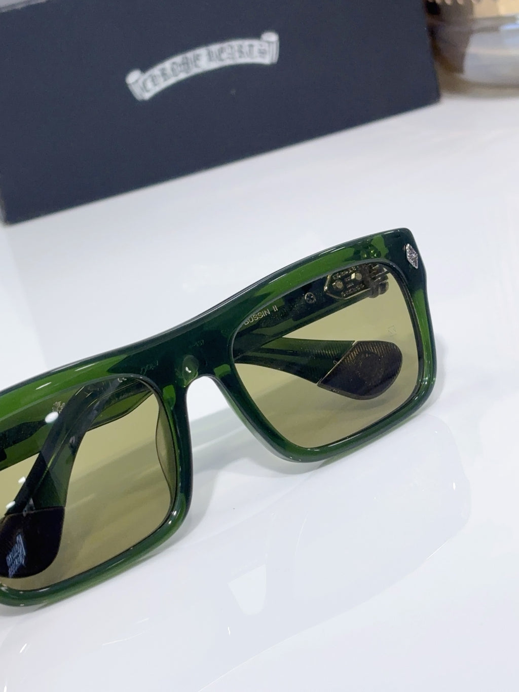 Chrome glasses close-up, green frames with tan-tinted lenses and temple logo