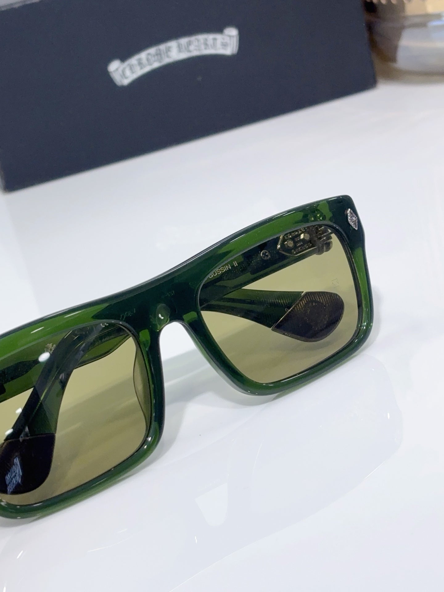 Chrome glasses close-up, green frames with tan-tinted lenses and temple logo