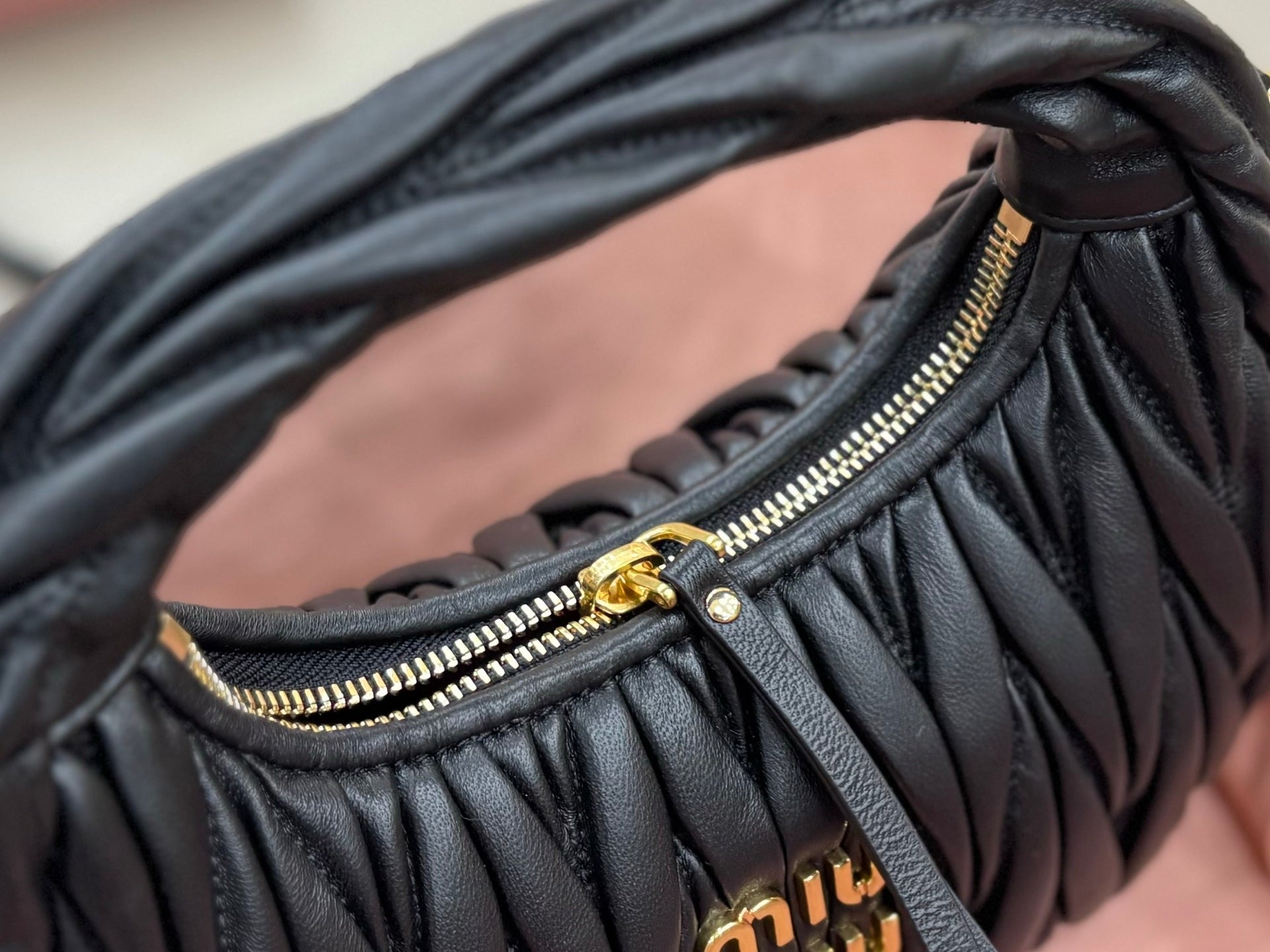Black leather handbag close-up with zip opening, gold zipper track, and ruched panel texture