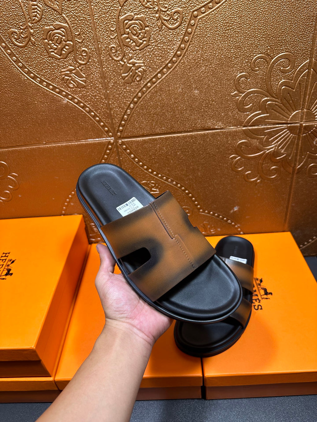 Hand holding Hermes slide sandals with gray-tan straps and black footbed