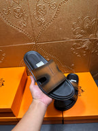 Hand holding Hermes slide sandals with gray-tan straps and black footbed