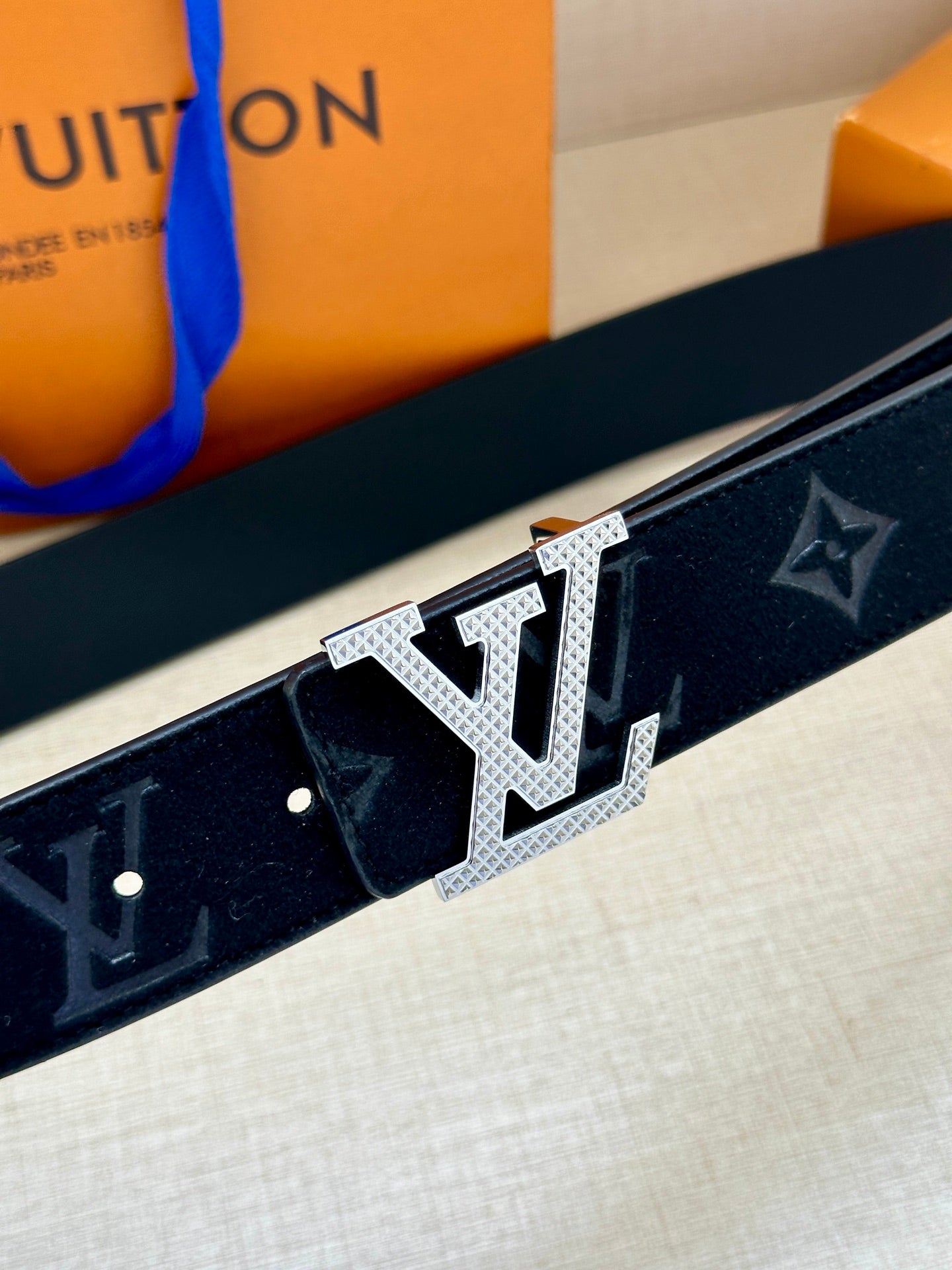Dark blue Louis-style monogram belt with close-up silver buckle and textured metal edges.