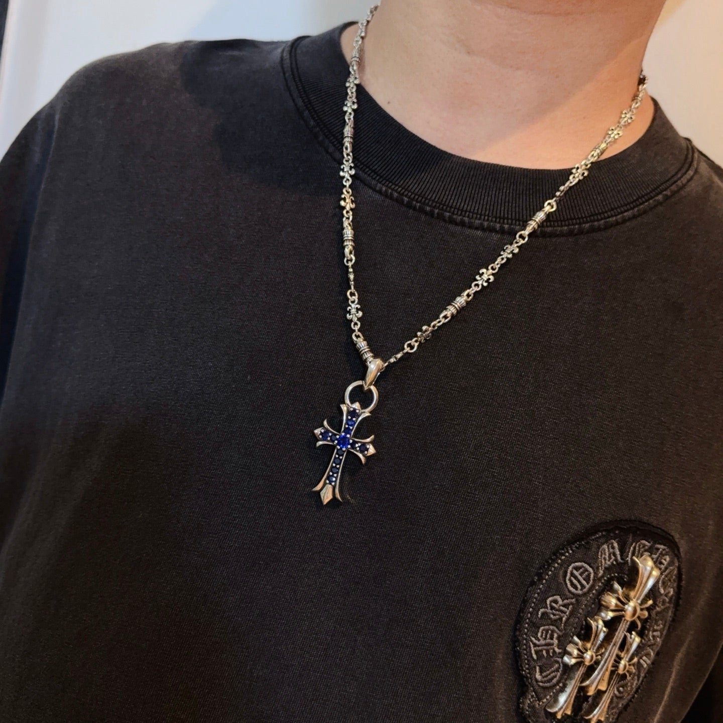 Silver cross pendant necklace with blue enamel charm on a dark crewneck shirt