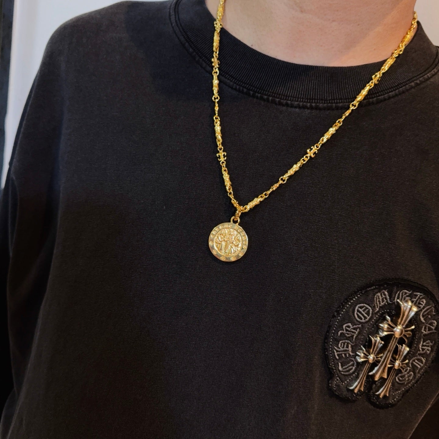 Gold-tone coin pendant necklace worn on dark crewneck shirt with patch