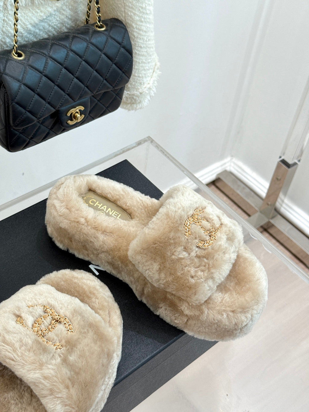 Beige faux fur slide sandals with gold logo detail, shown on display box