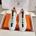 White low-top sneakers with black side straps and orange laces, chunky rubber sole