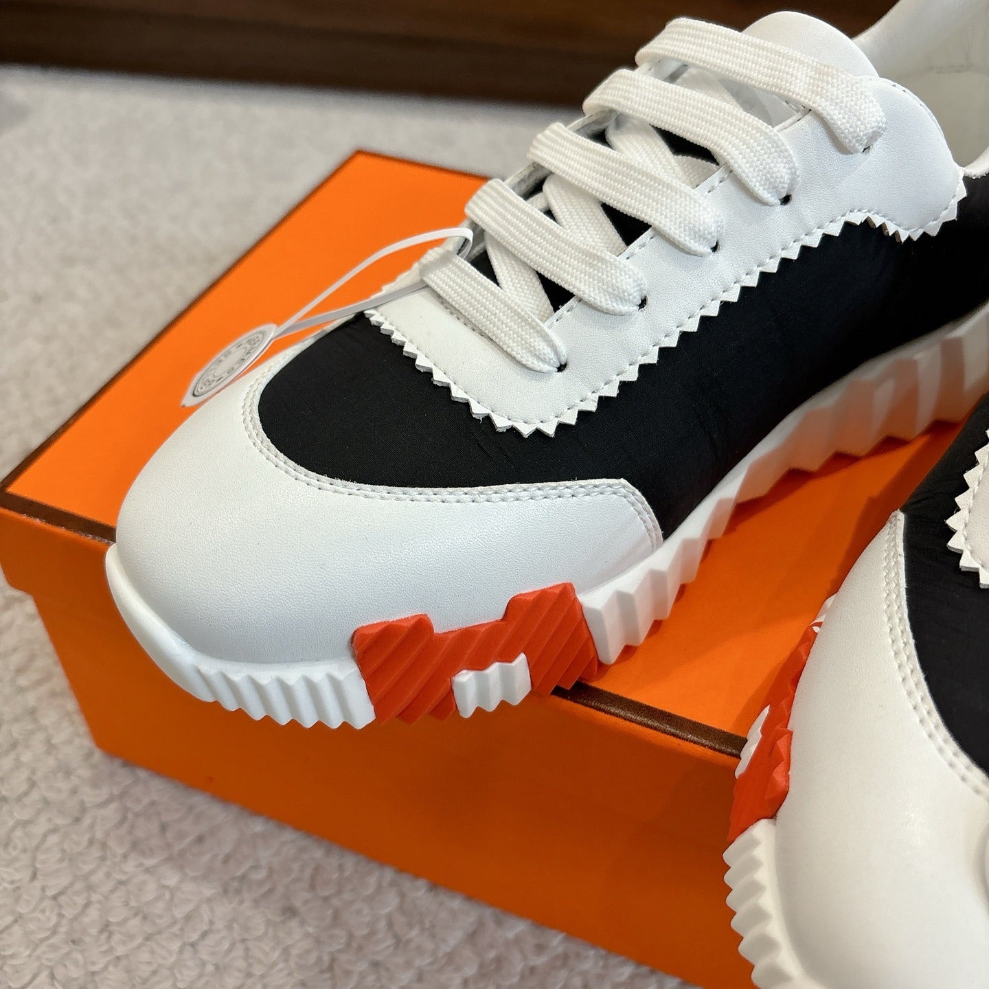 Hermes unisex sneaker toe, white leather with black side panel and orange-red sole trim.
