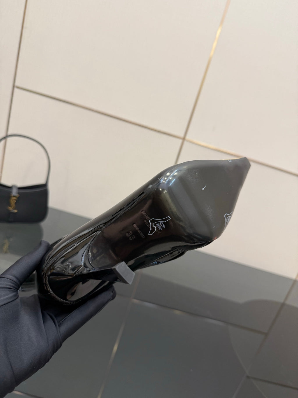 Black patent ankle boot held in gray-gloved hand, side view of pointed gold heel