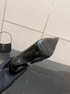 Black patent ankle boot held in gray-gloved hand, side view of pointed gold heel