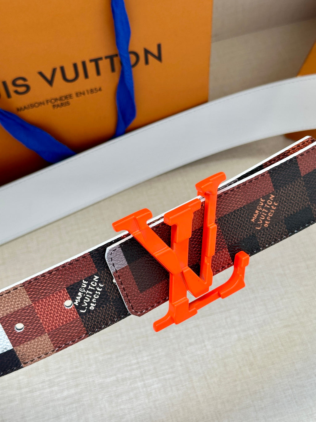 Louis Vuitton belt buckle close-up on dark checkered strap with orange LV letters