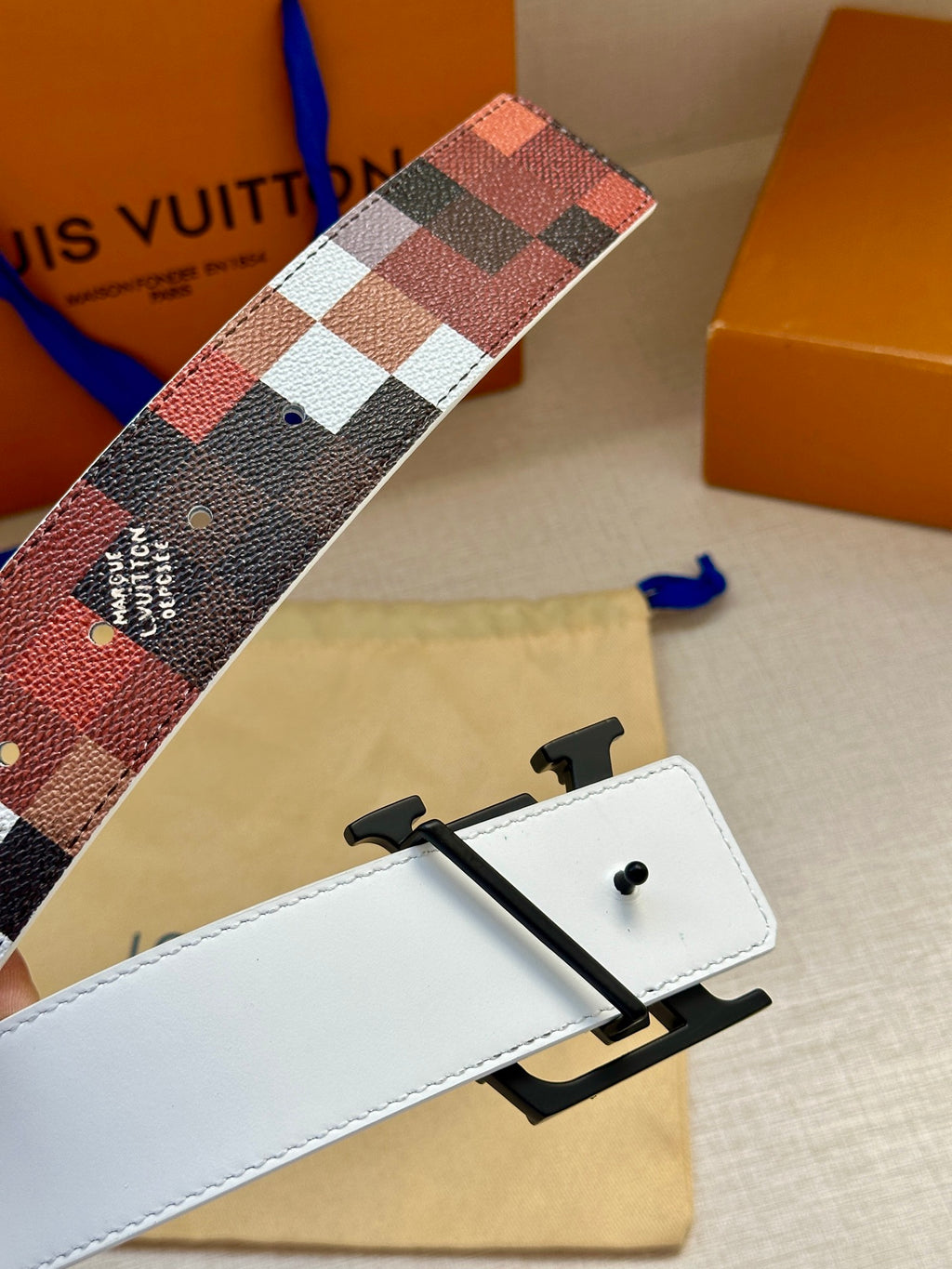 Louis Vuitton belt detail with white leather strap and black buckle over check pattern.
