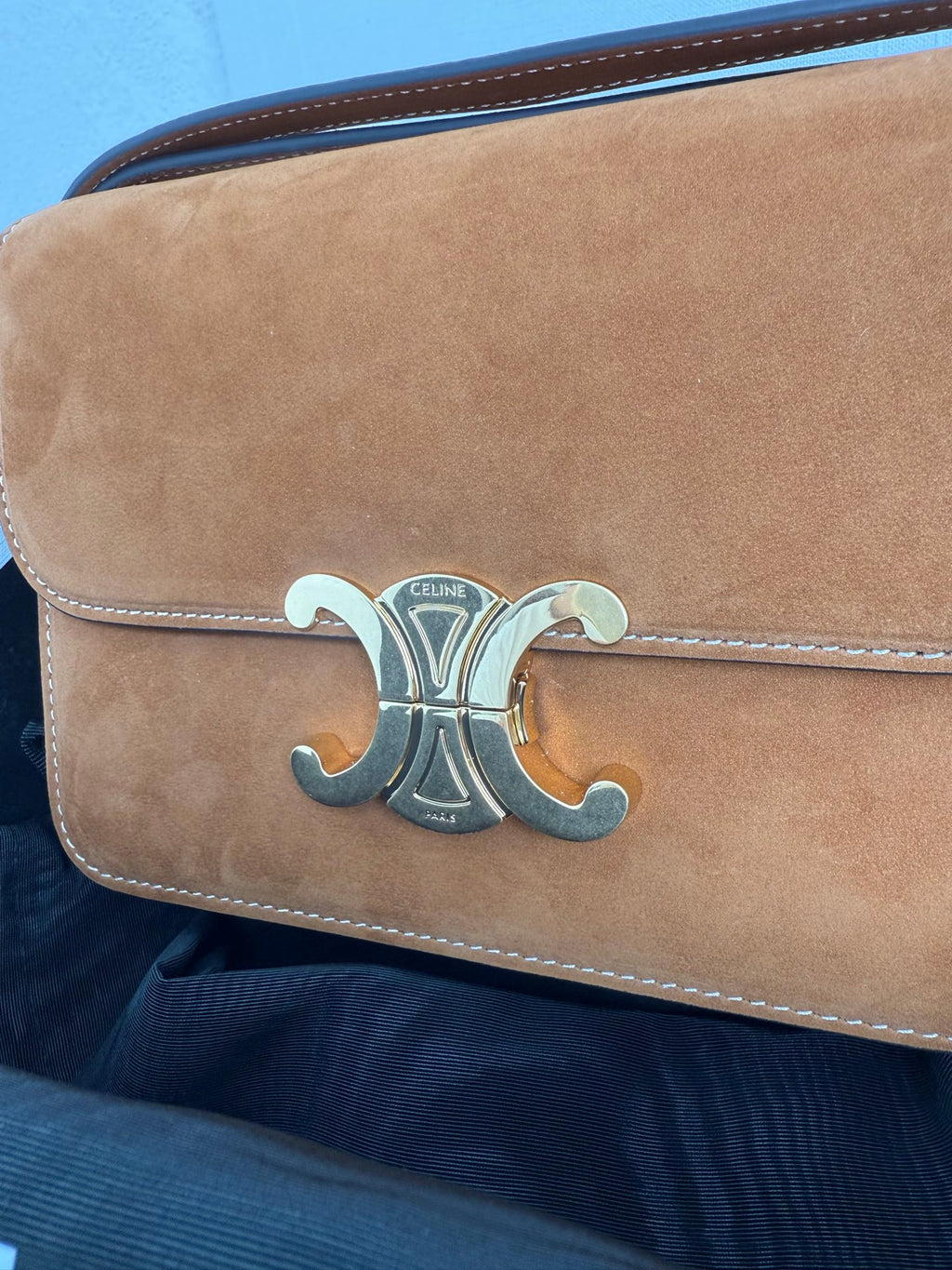 Tan suede bag flap with gold-tone metal emblem and stitched edge trim.