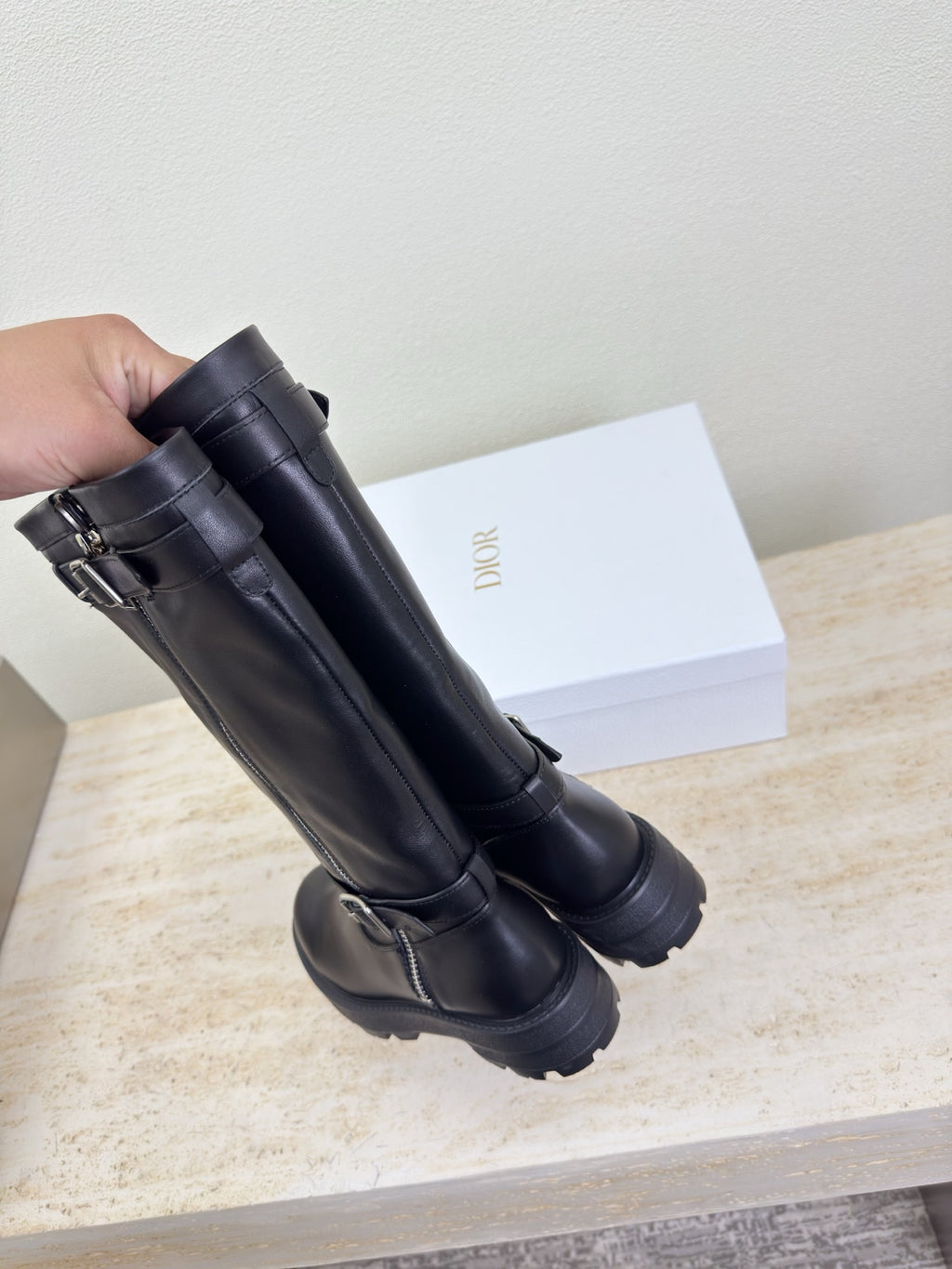 Hand holding black knee-high boots with buckles and side zipper, close view