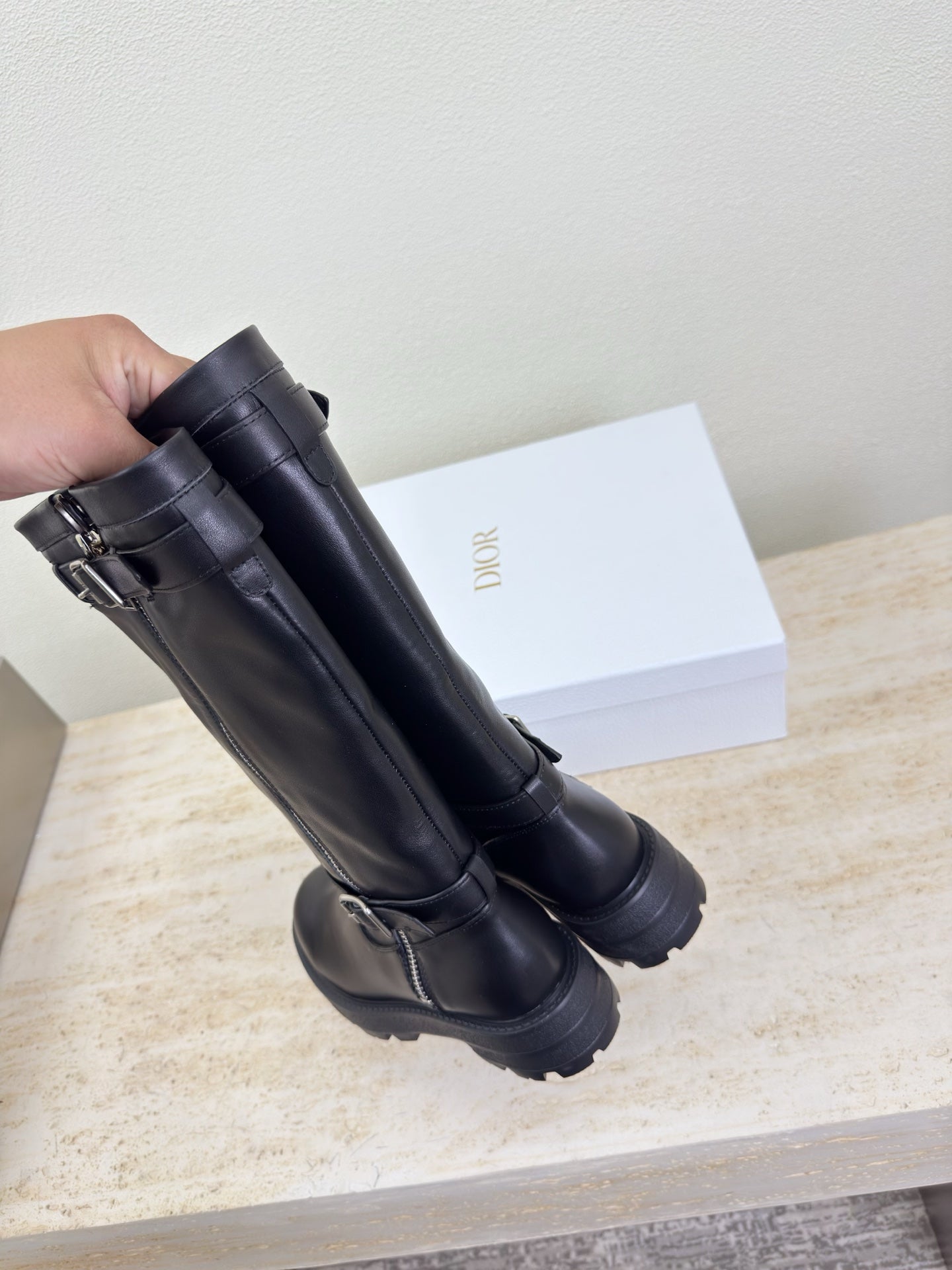 Hand holding black knee-high boots with buckles and side zipper, close view