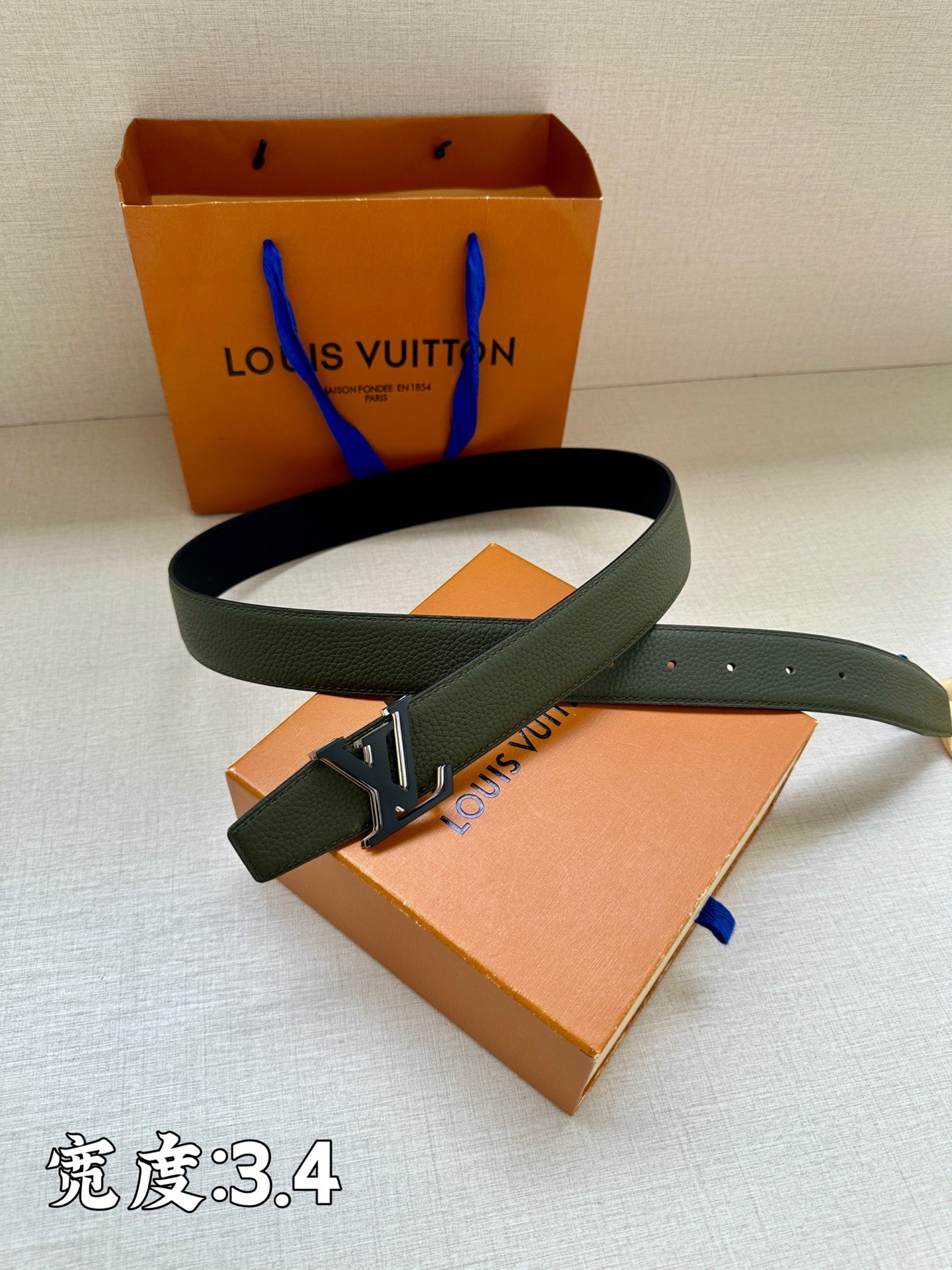 Louis Vuitton leather belt with dark green strap and silver LV buckle, flat on box.