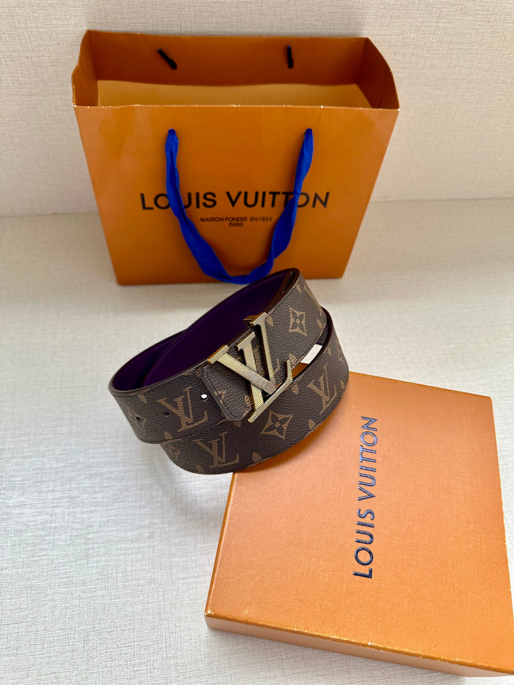 Louis Vuitton belt with dark monogram strap and gold LV buckle, laid across box.