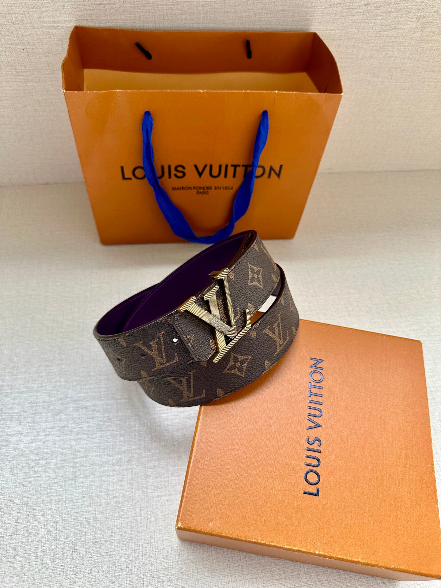 Louis Vuitton belt with dark monogram strap and gold LV buckle, laid across box.