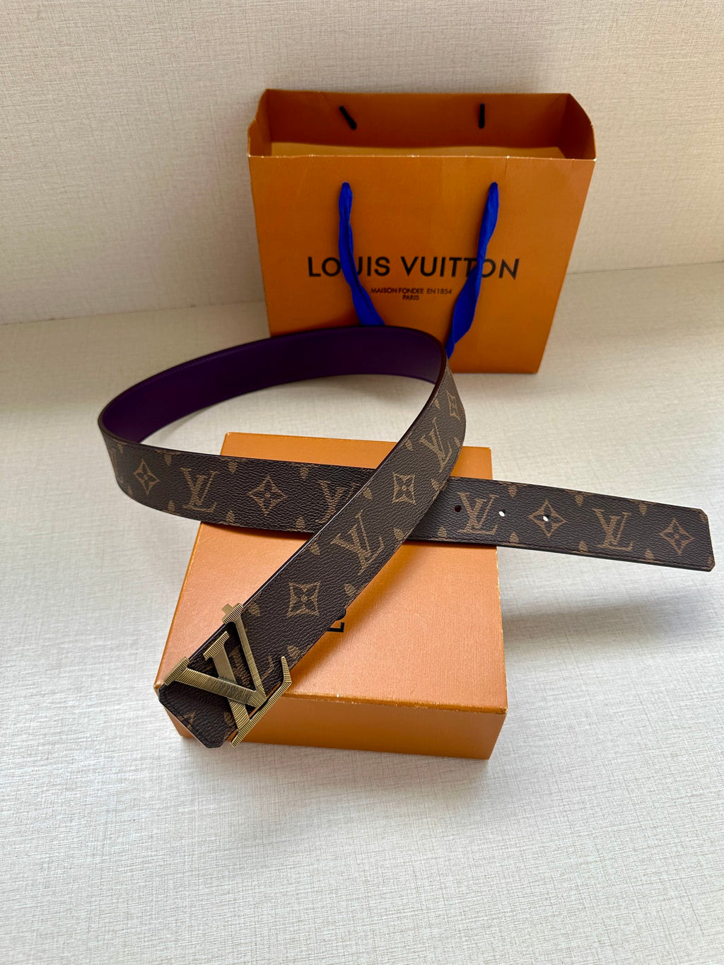 Louis Vuitton belt laid flat, dark monogram strap with gold LV buckle and purple underside.