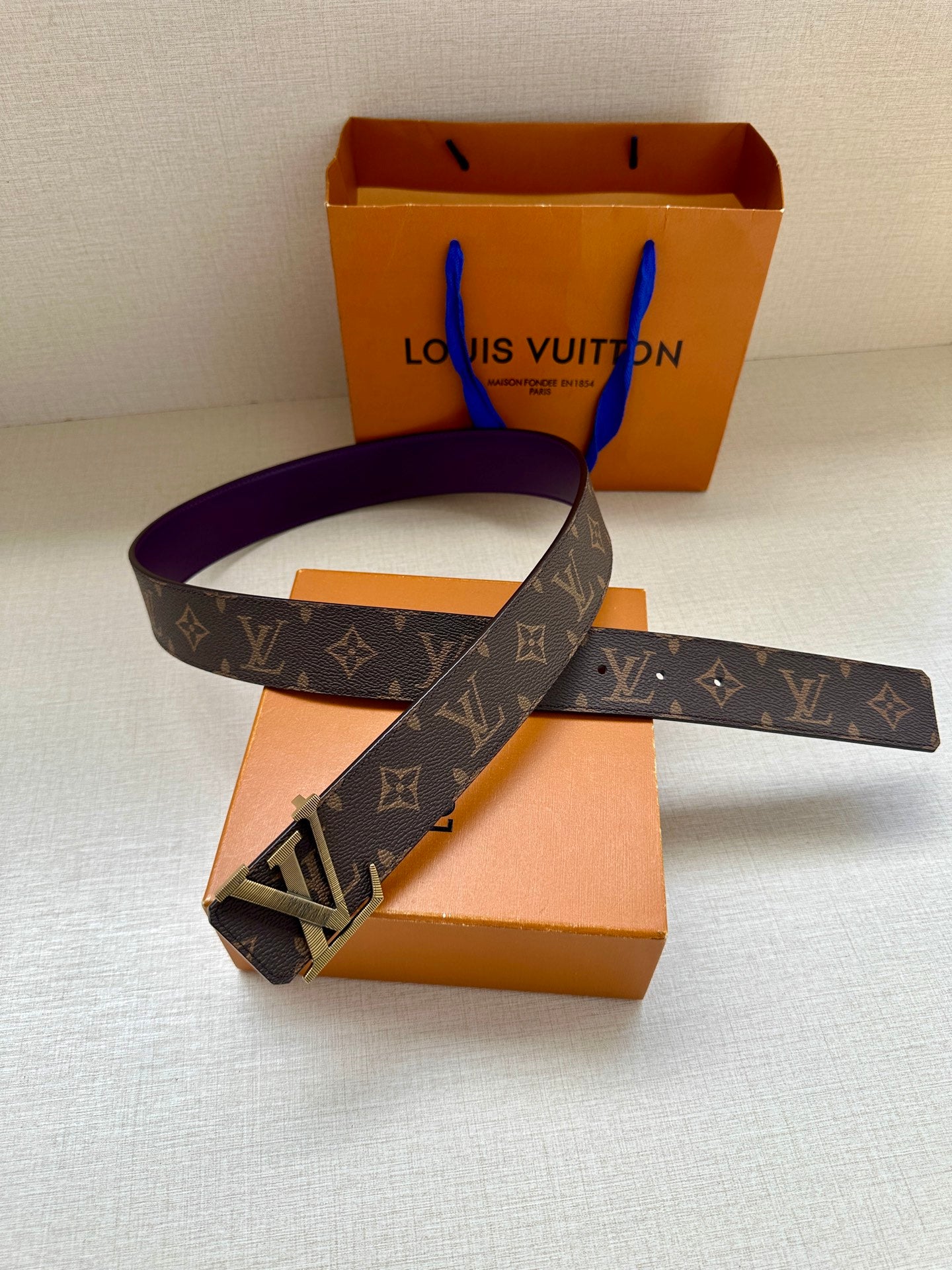 Louis Vuitton belt laid flat, dark monogram strap with gold LV buckle and purple underside.