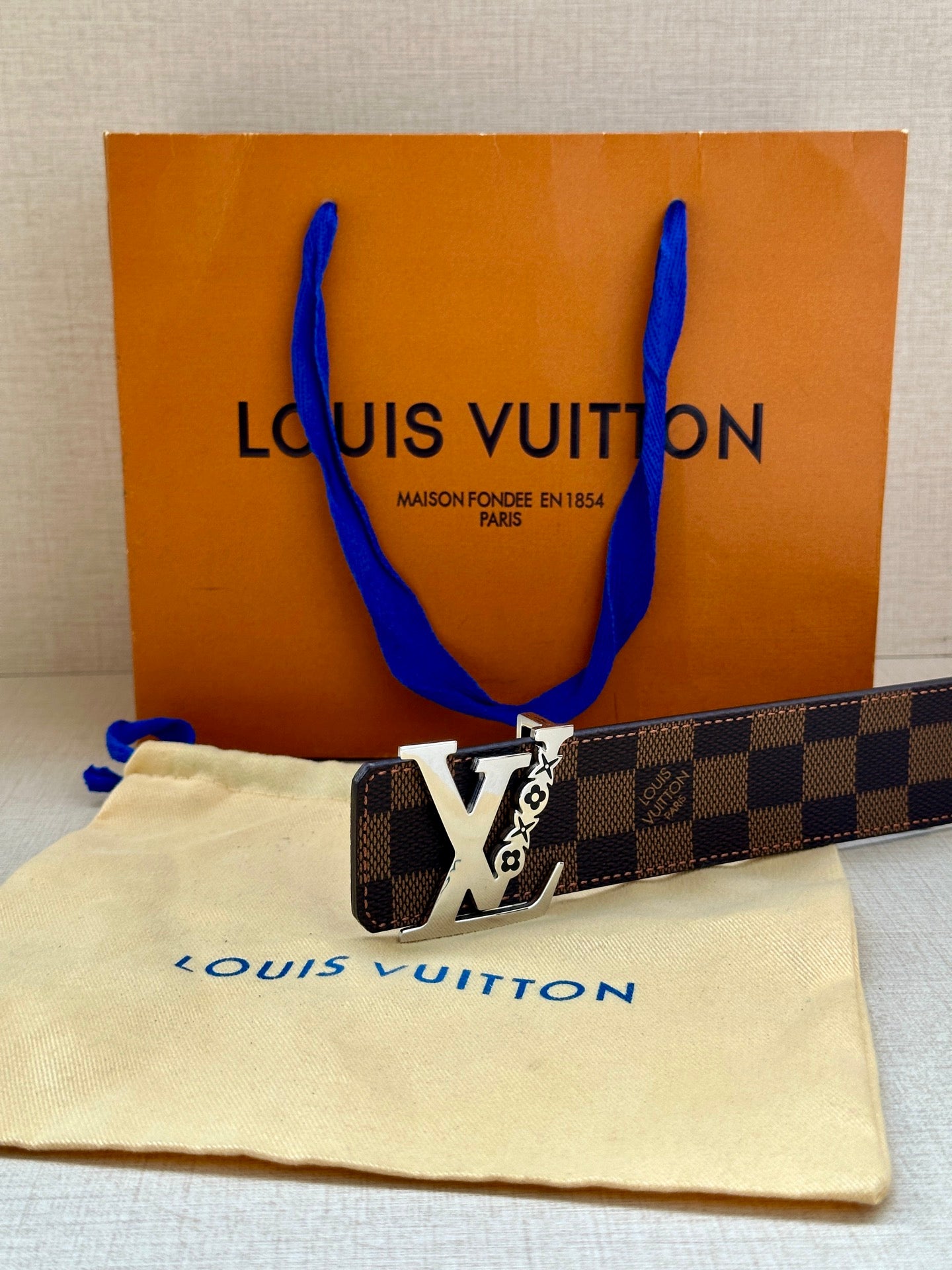 Louis Vuitton belt with dark check strap and gold LV buckle, lying beside logo dust bag