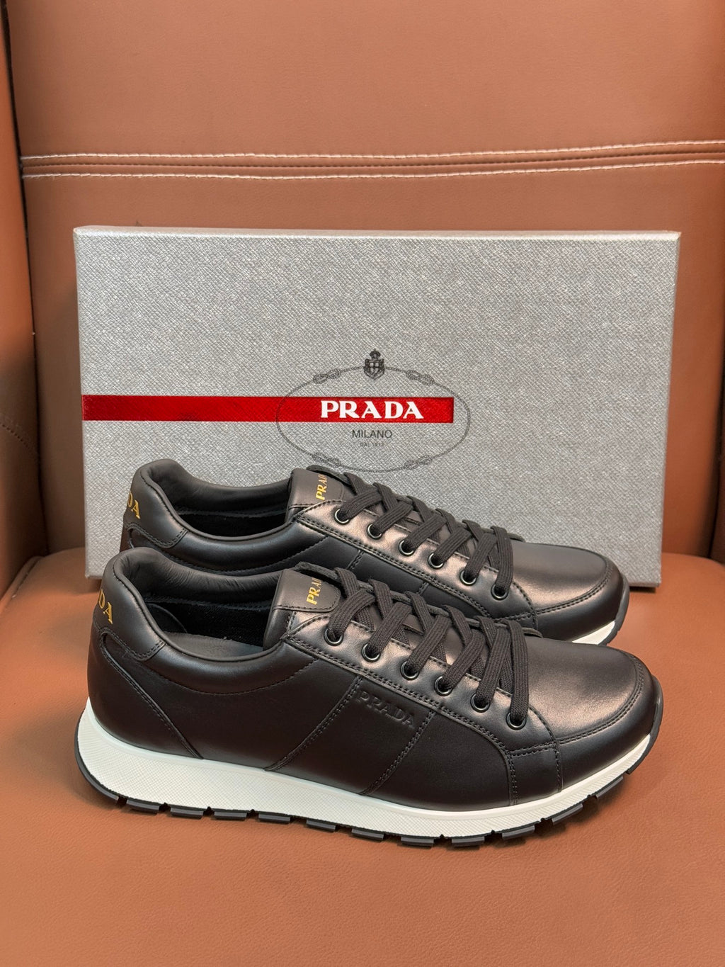 Prada unisex sneakers pair on the box, dark leather lace-up with white midsole trim
