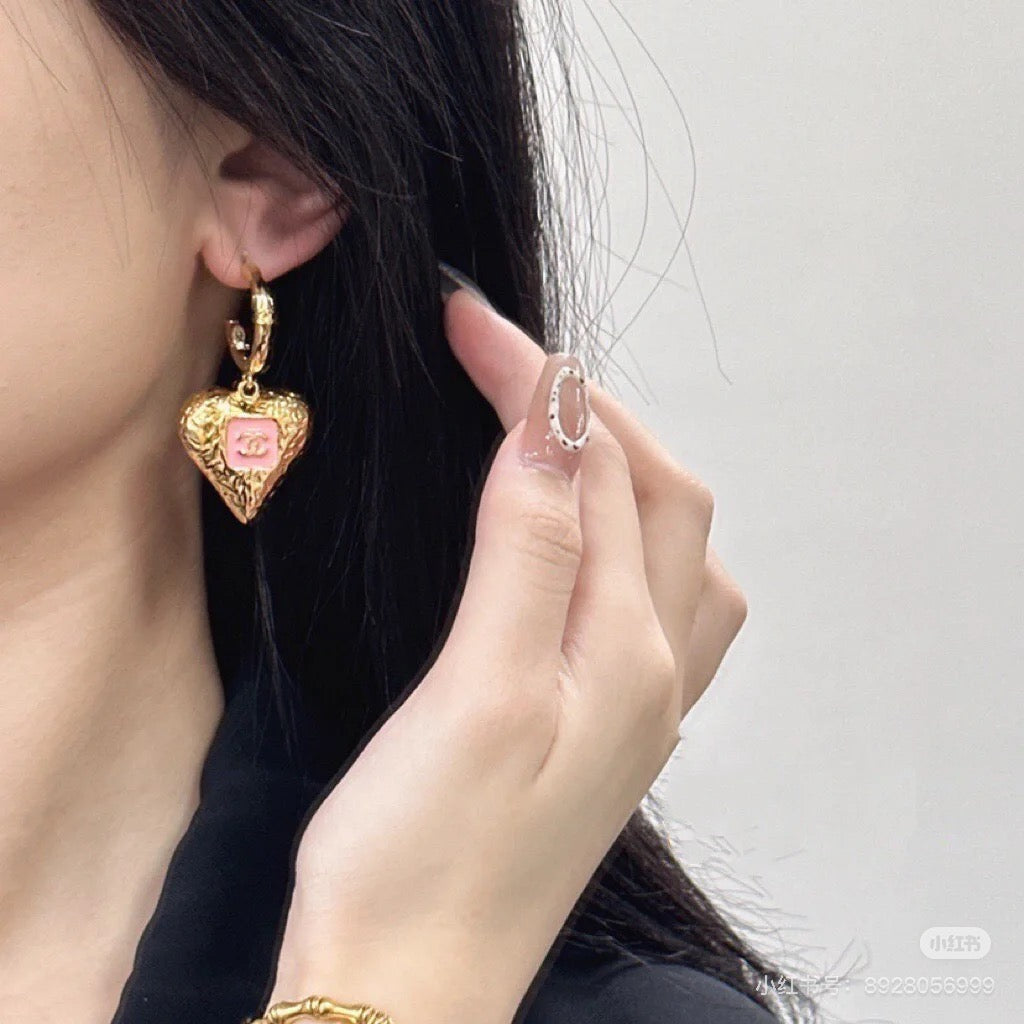 Gold-tone heart drop earrings with pink enamel logo on model's ear.