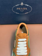 Unisex Prada sneaker close-up, tan suede upper with green side panel and white lace.