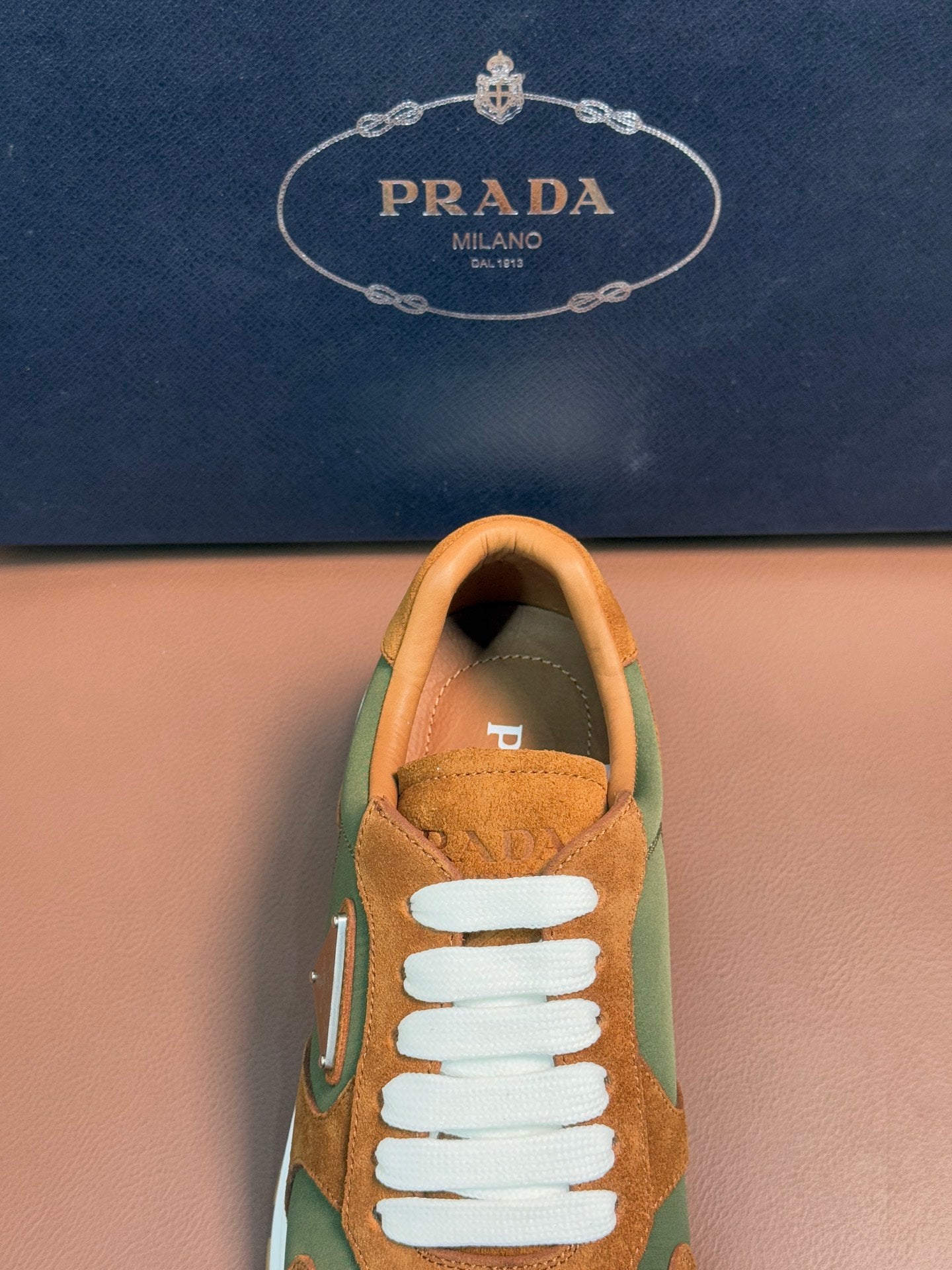 Unisex Prada sneaker close-up, tan suede upper with green side panel and white lace.