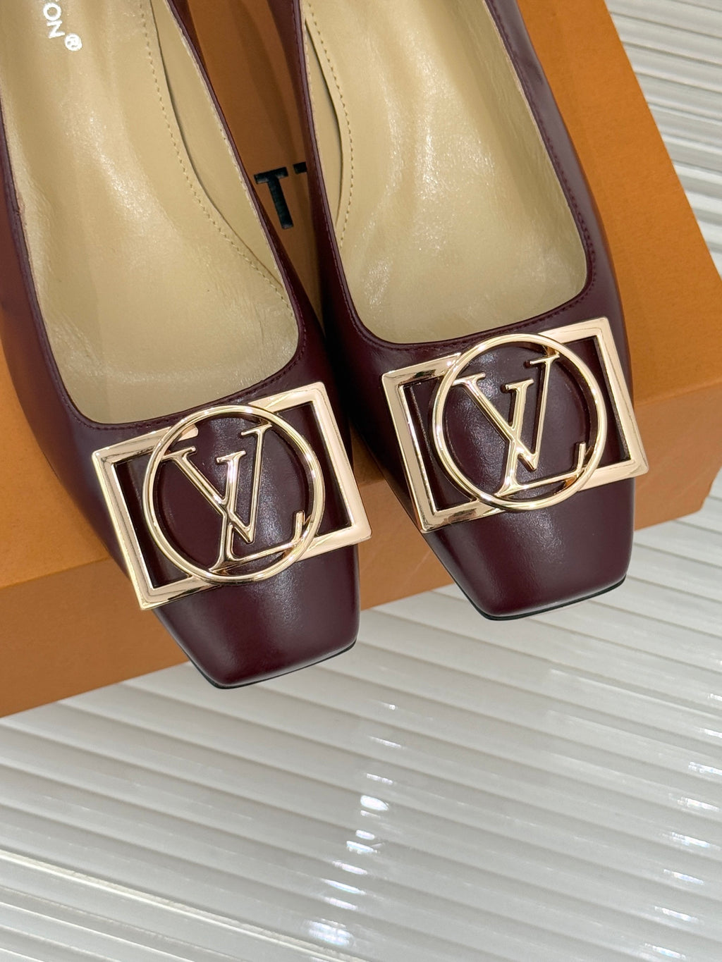 Close-up of burgundy low-heel pumps with gold square logo buckle detail.