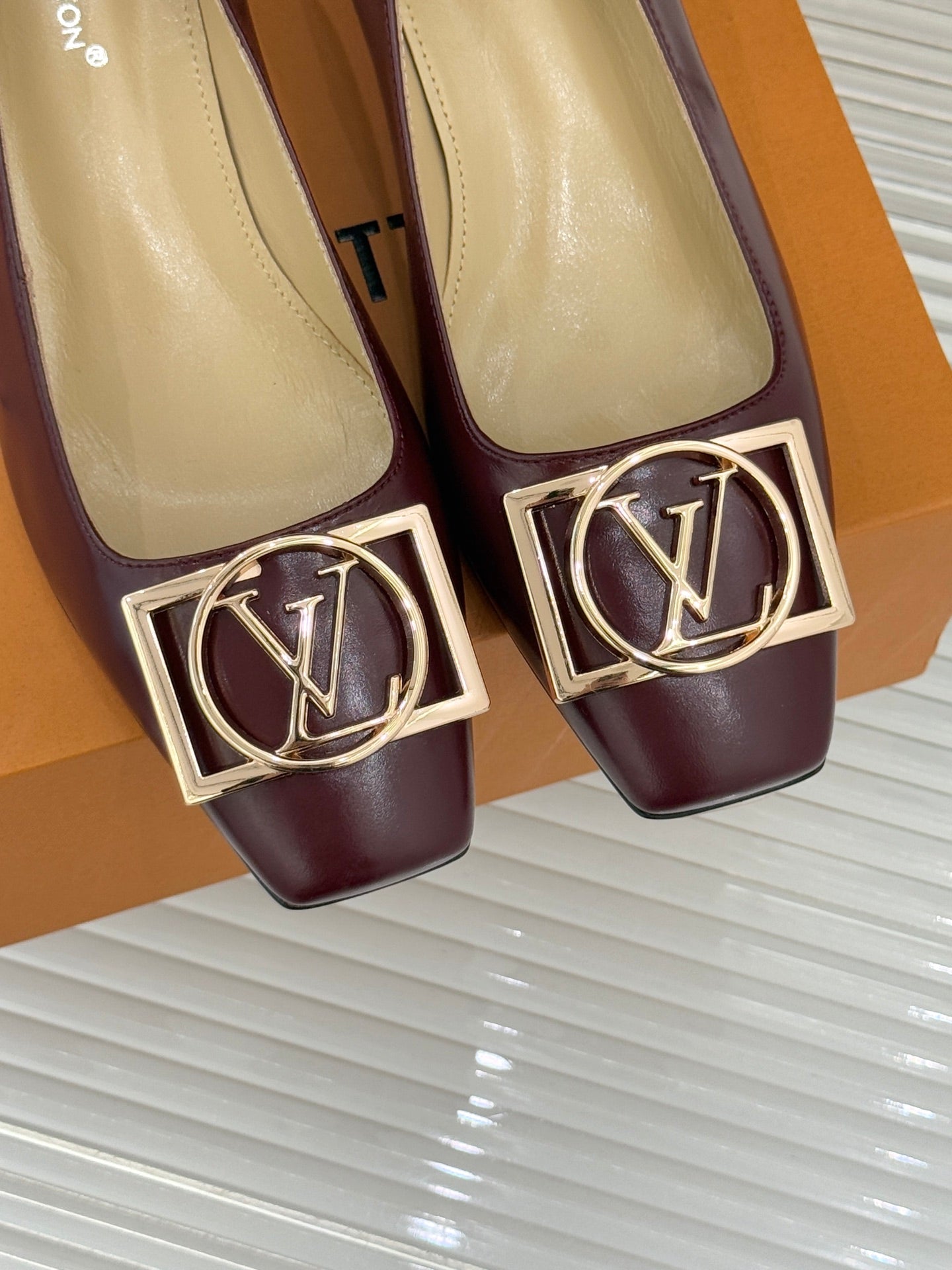 Close-up of burgundy low-heel pumps with gold square logo buckle detail.