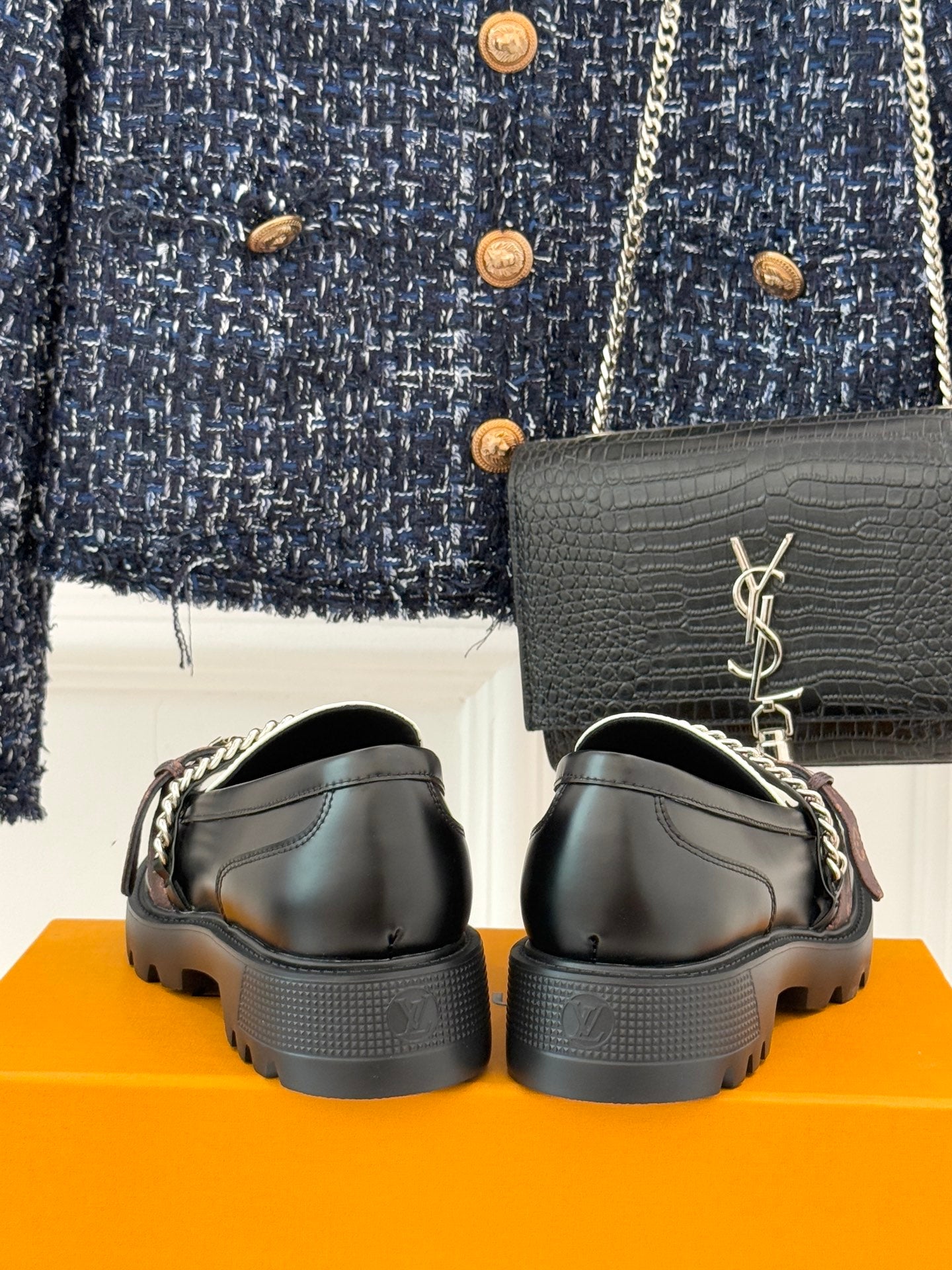 Back view of black loafers on orange box, with chunky sole and metal logo charm