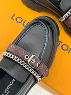 Black leather loafer with brown monogram strap, metal eyelets, and chain trim