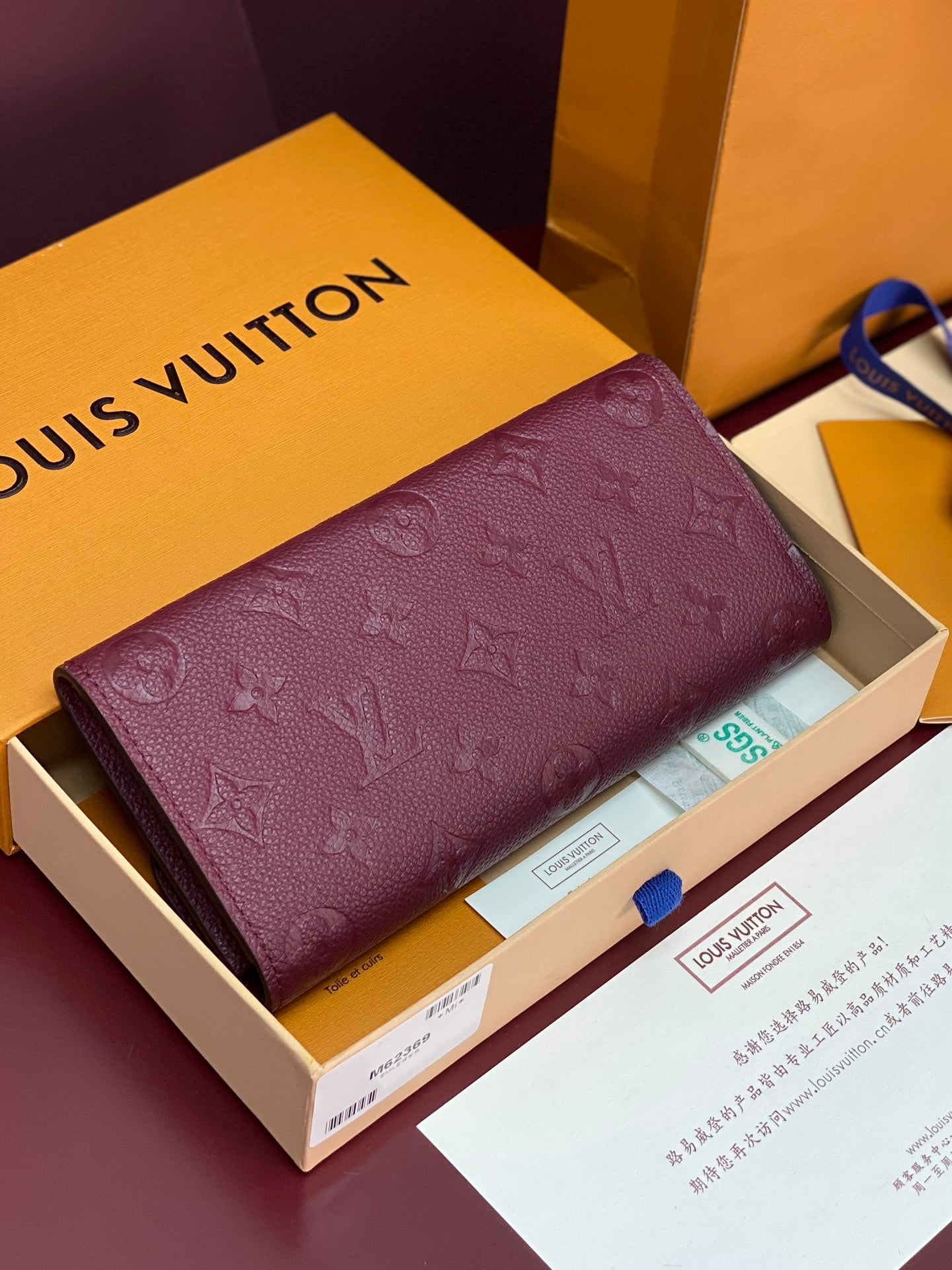 Small bag with long flap and burgundy/purple embossed monogram pattern, open box view.
