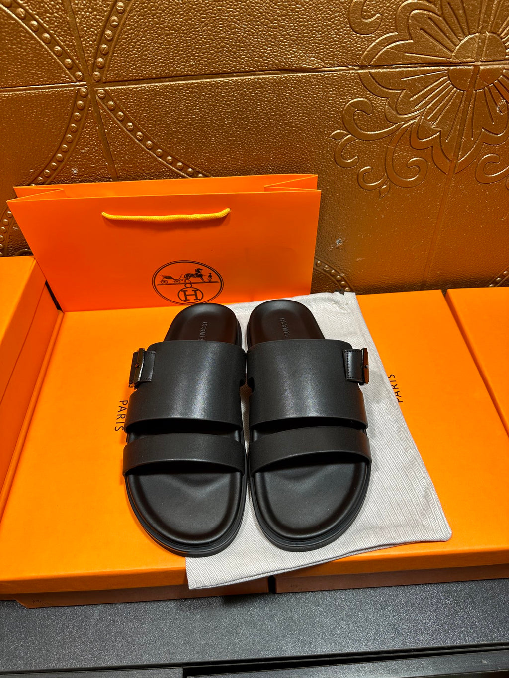 Black leather slide sandals, two wide straps with buckle, on dust bag
