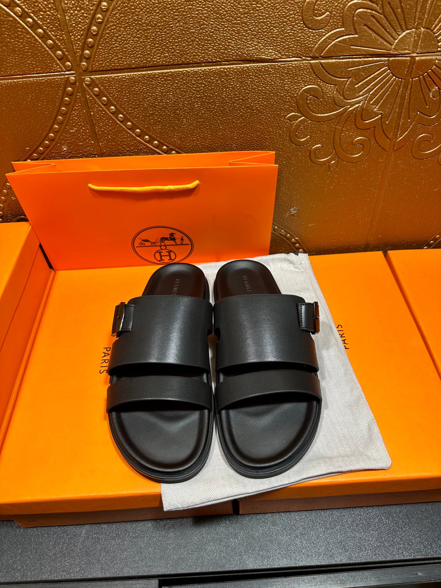 Black leather slide sandals, two wide straps with buckle, on dust bag