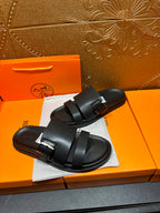 Black leather slide sandals, angled view of wide buckled straps on dust bag
