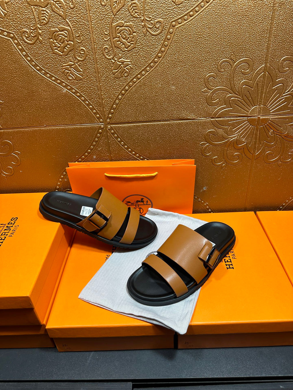 Tan and black Hermes slide sandals held in hand over orange box