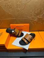 Tan and black Hermes slide sandals held in hand over orange box