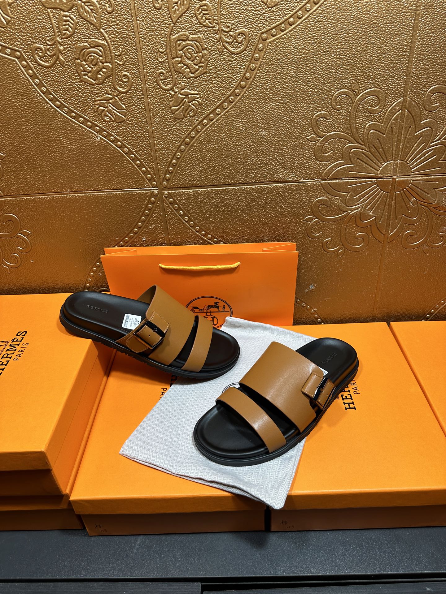 Tan and black Hermes slide sandals held in hand over orange box